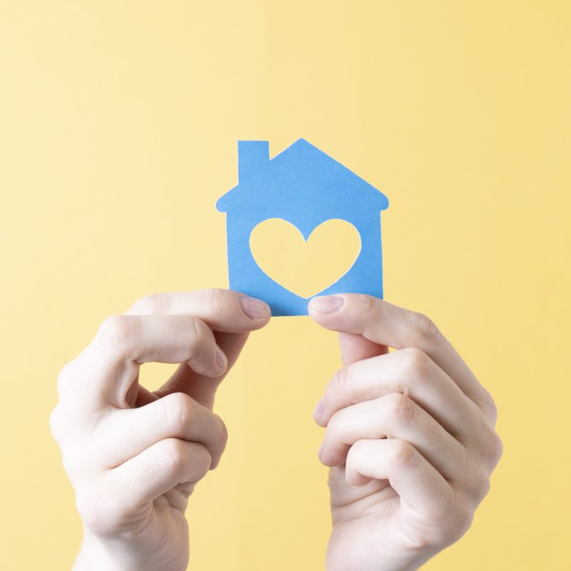 blue paper house with heart cut out in female hands on yellow background illustrating you can sell your home easily with kindness